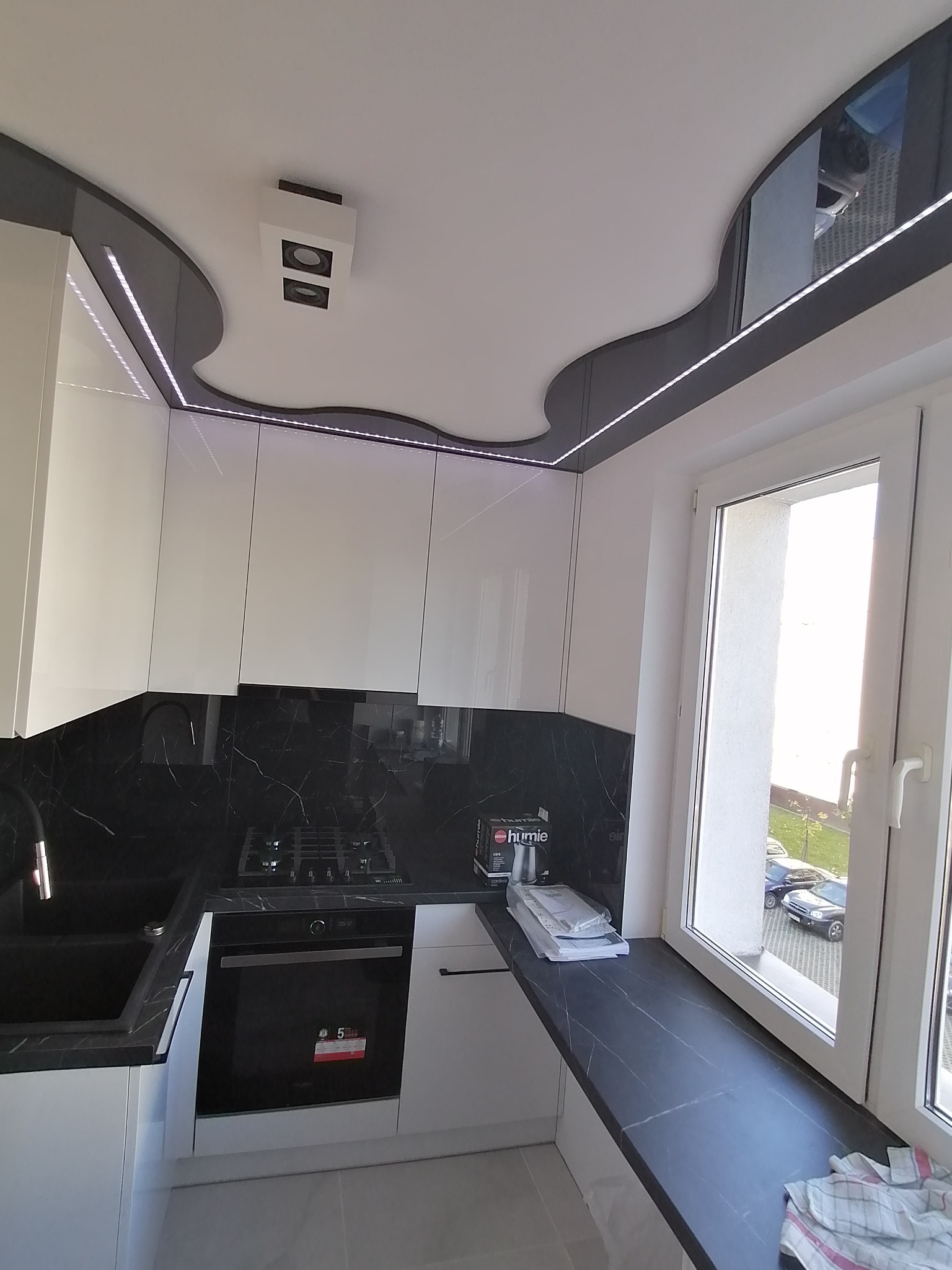 Nowoczesna kuchnia z białymi szafkami, czarnym blatem i marmurowym backsplashiem, oświetleniem LED pod sufitem i oknem z widokiem na zewnątrz.