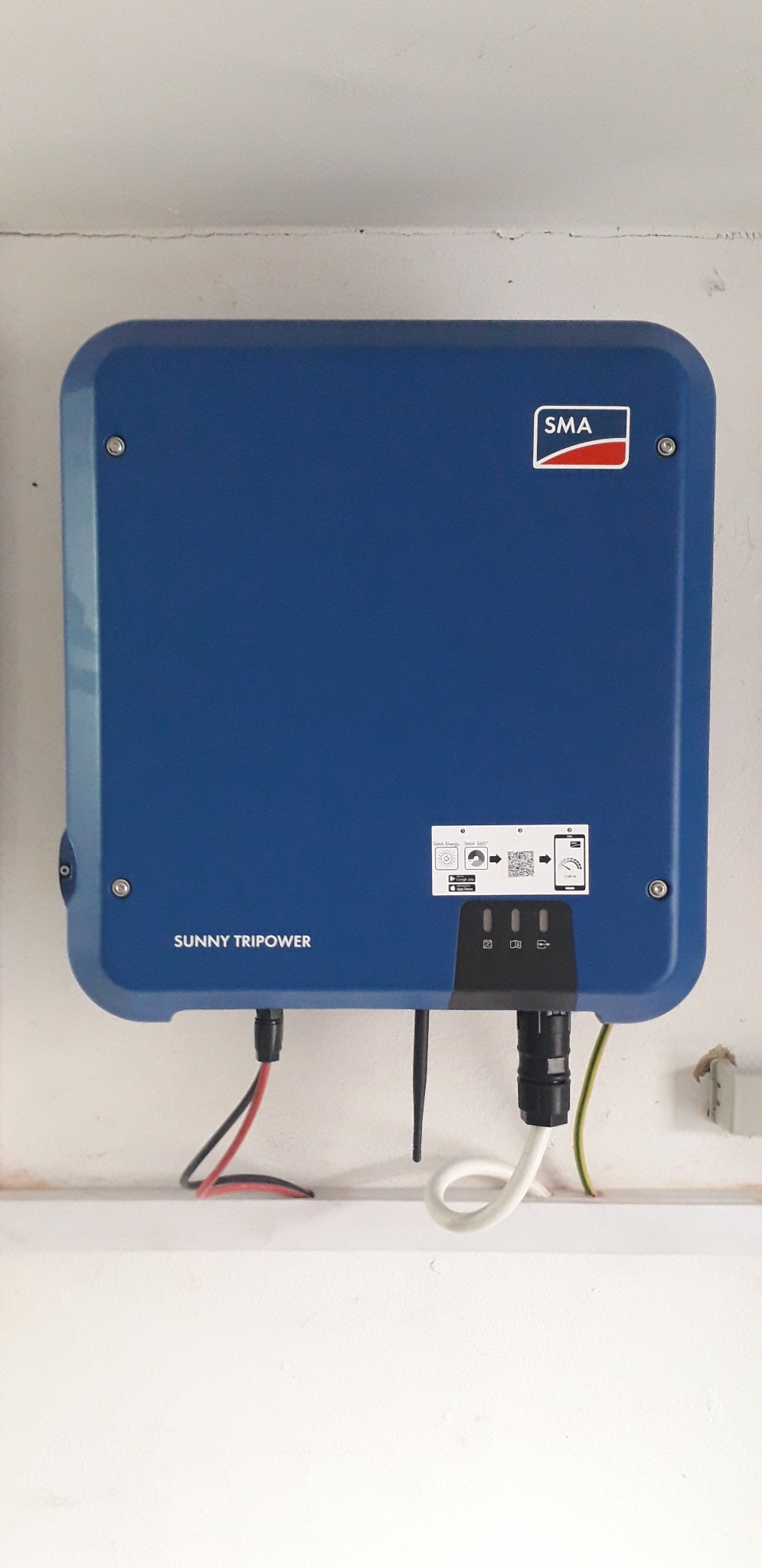Niebieski falownik SMA Sunny Tripower zamontowany na białej ścianie, z widocznymi kablami i naklejką z kodem QR.