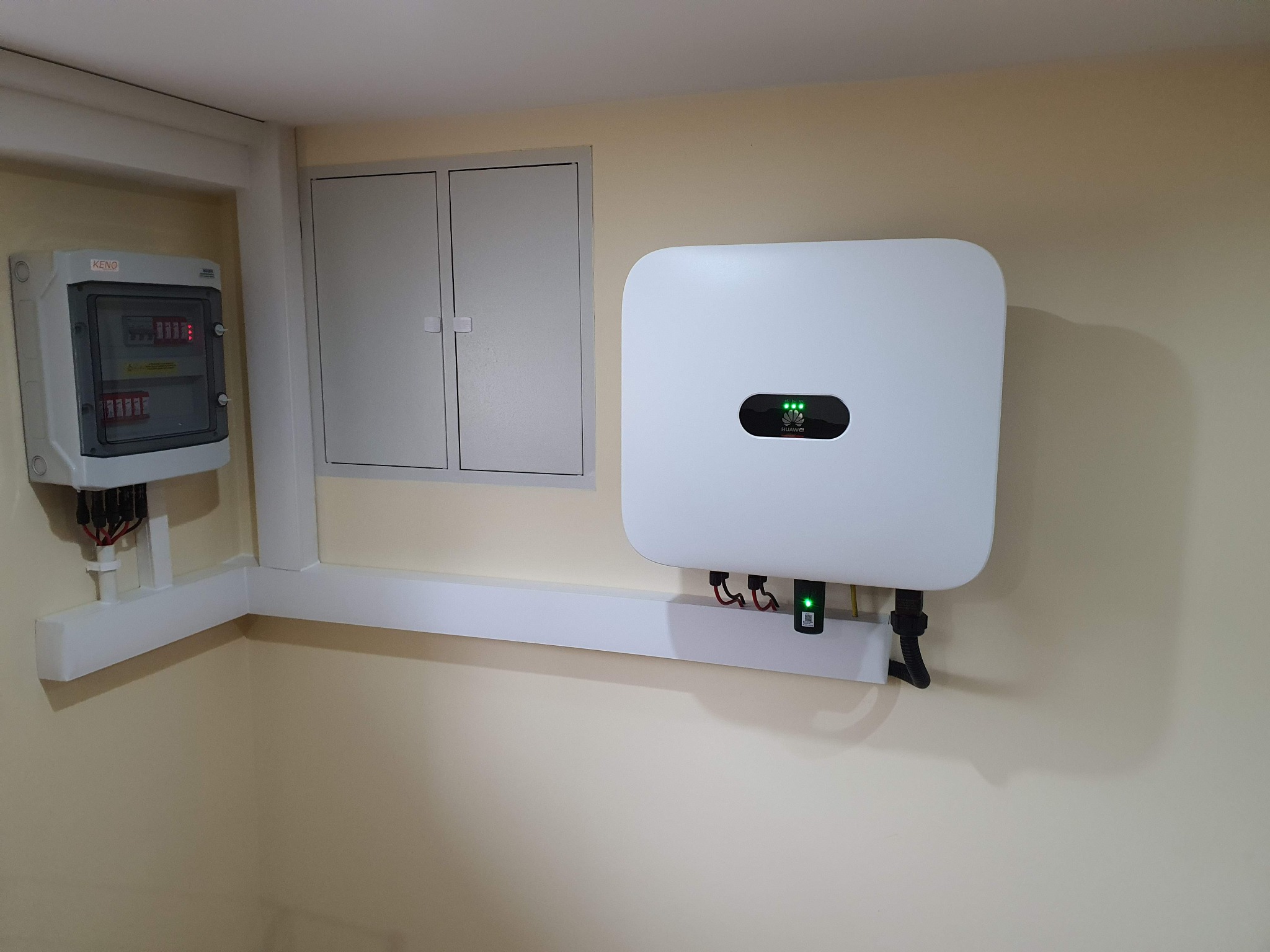 Biały inwerter fotowoltaiczny Huawei zamontowany na ścianie wewnątrz pomieszczenia, obok skrzynka elektryczna KENO z widocznymi bezpiecznikami.