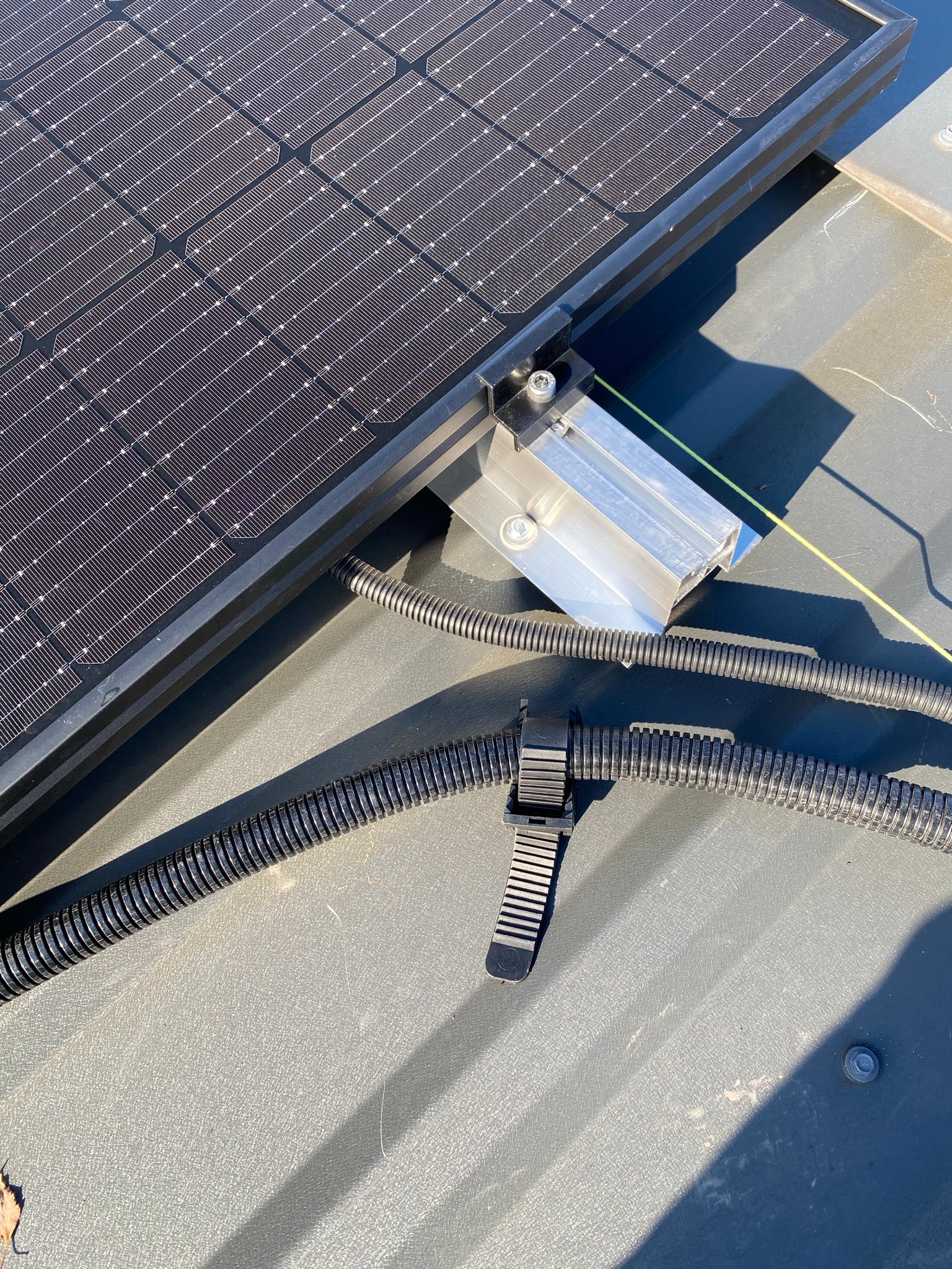 Panel słoneczny przymocowany do szarego dachu za pomocą aluminiowego wspornika; widoczne czarne kable w karbowanej osłonie, przymocowane plastikową opaską zaciskową.