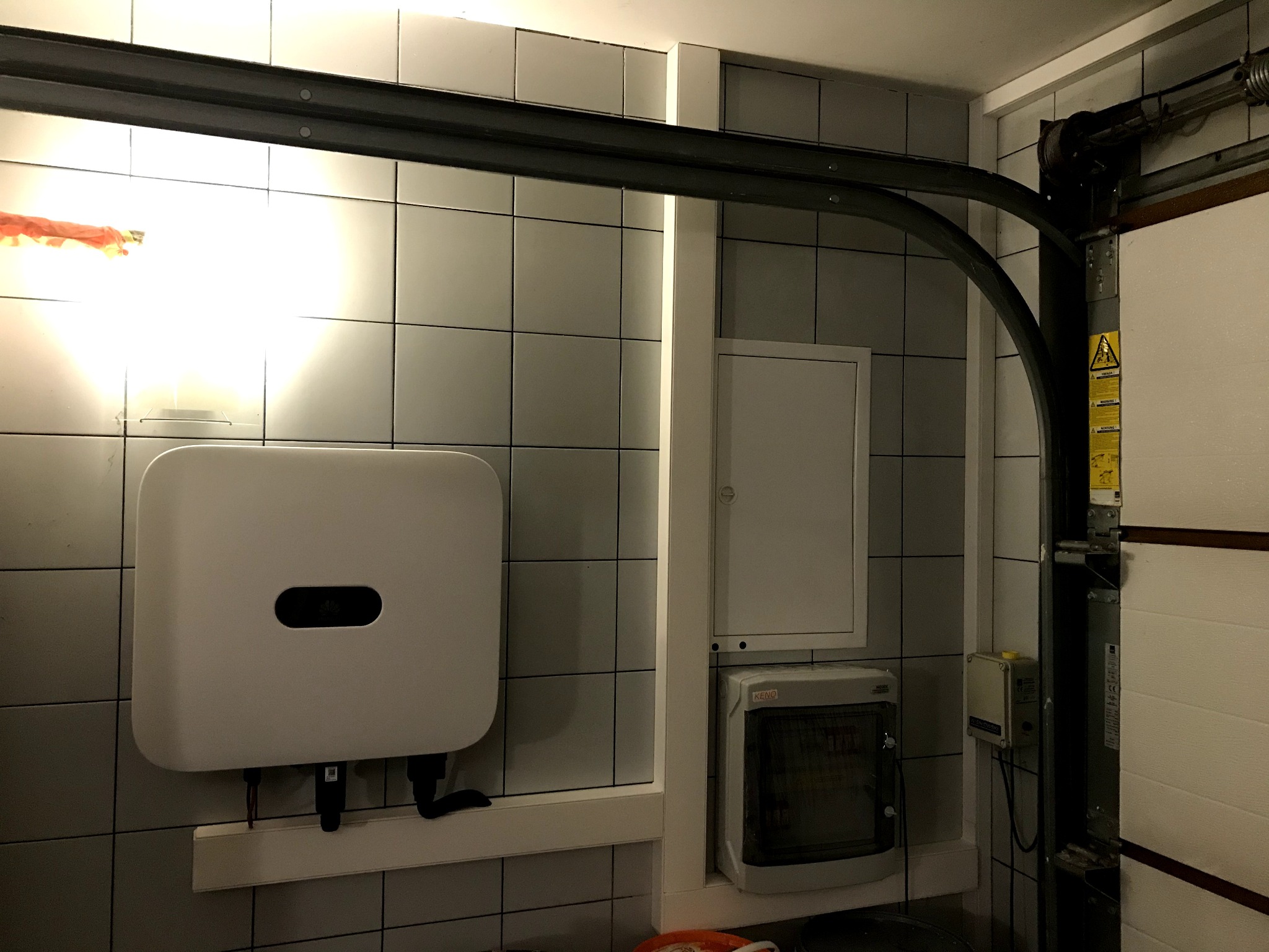 Biały falownik fotowoltaiczny Huawei zamontowany na ścianie z szarych płytek w garażu, widoczne przewody i elementy instalacji elektrycznej.