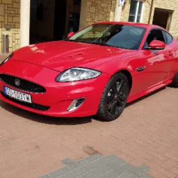 Czerwony Jaguar XK zaparkowany przed budynkiem warsztatu z cegły, widoczny numer rejestracyjny GD 103TW i czarne felgi.