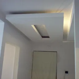 Nowoczesny sufit podwieszany z prostokątną wnęką oświetleniową, białe ściany w tle, widoczne drzwi zabezpieczone folią malarską.