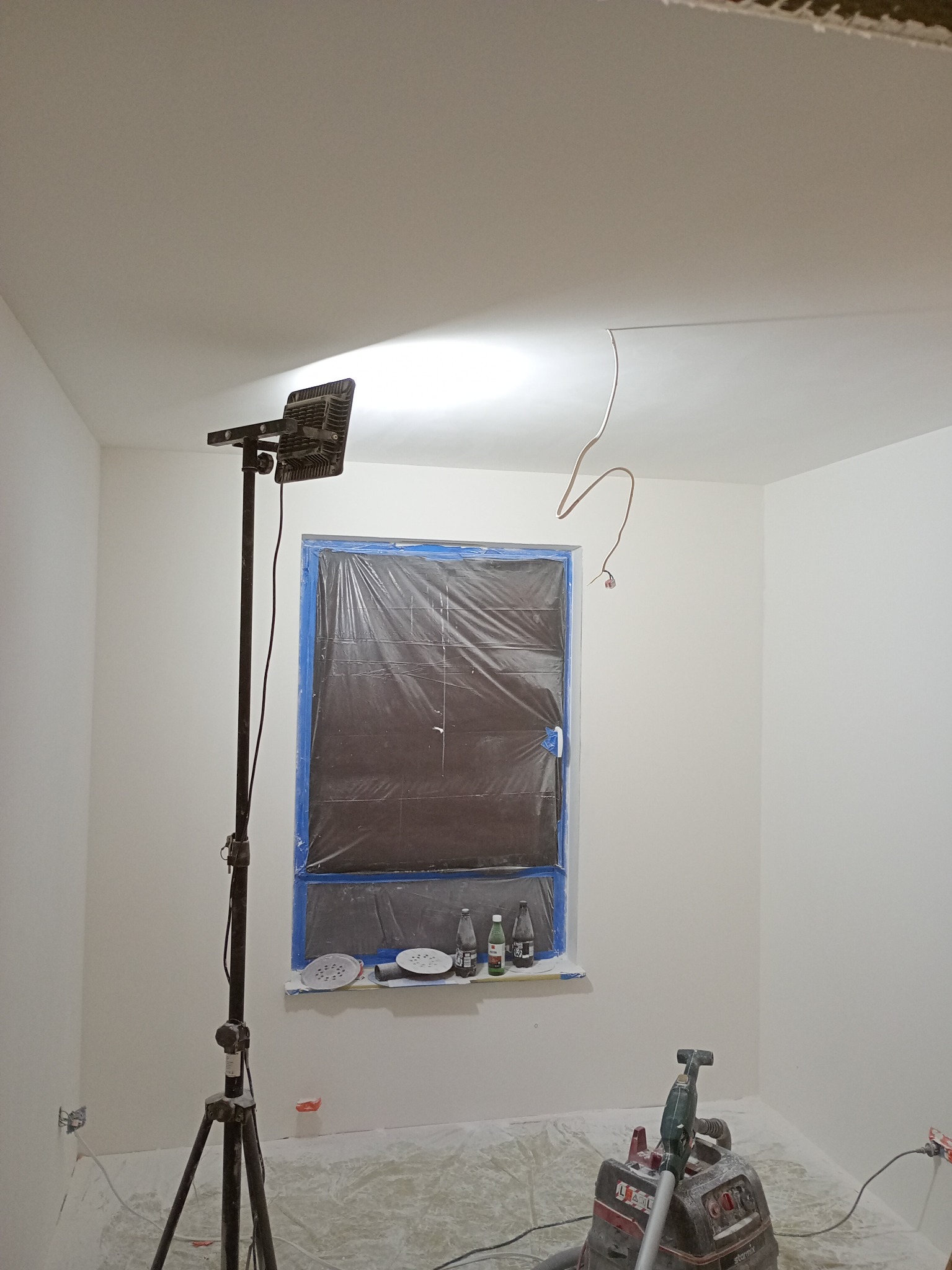 Białe ściany po szpachlowaniu, okno zabezpieczone folią, lampa na statywie i szlifierka do gładzi. Widoczne ślady pyłu po szlifowaniu. Przygotowanie do malowania.