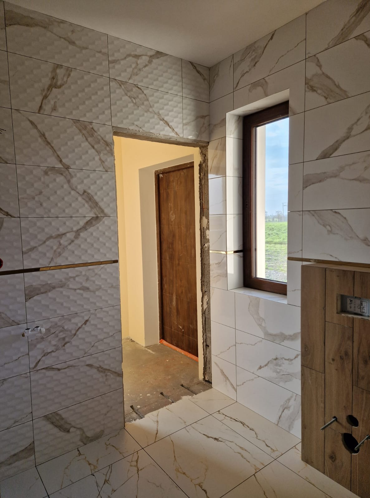 Łazienka w trakcie wykańczania: białe płytki imitujące marmur z brązowymi żyłami, widoczne okno i drzwi. Częściowo ułożona podłoga i ściany z dekoracyjnymi listwami.