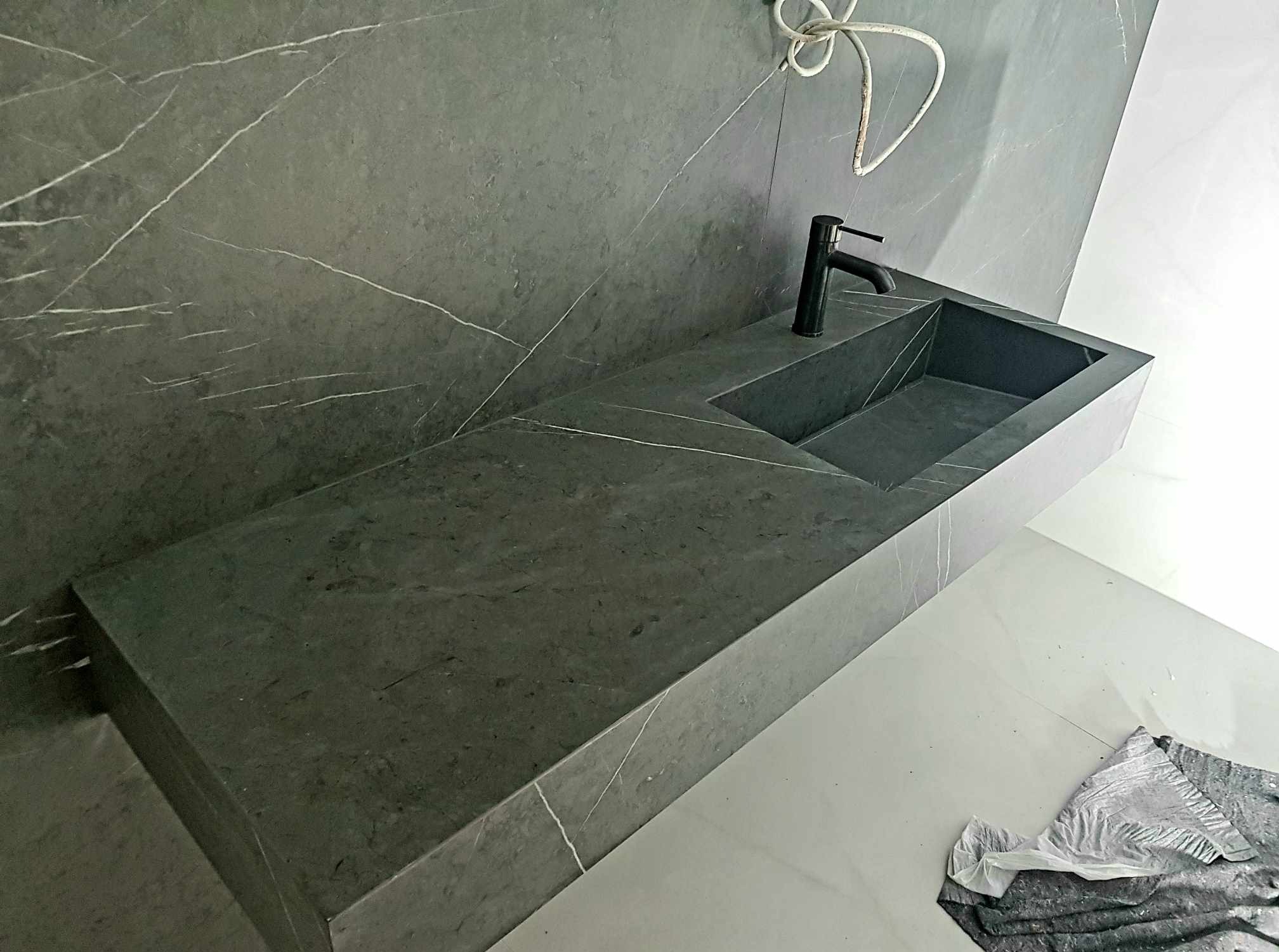 Nowoczesna łazienka z umywalką z szarego marmuru z czarną armaturą. Minimalistyczny design, widoczne białe żyłki na kamieniu. Częściowo widoczna instalacja.