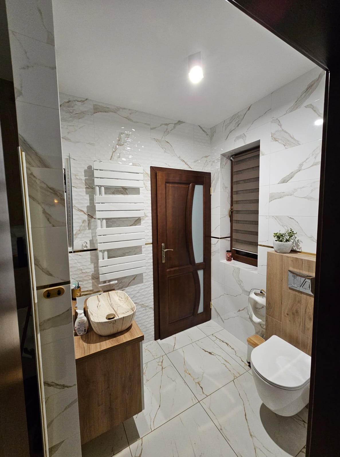 Nowoczesna łazienka z marmurowymi płytkami, drewnianą szafką pod umywalką i grzejnikiem drabinkowym. Uwagę zwraca nietypowa umywalka w odcieniach beżu.