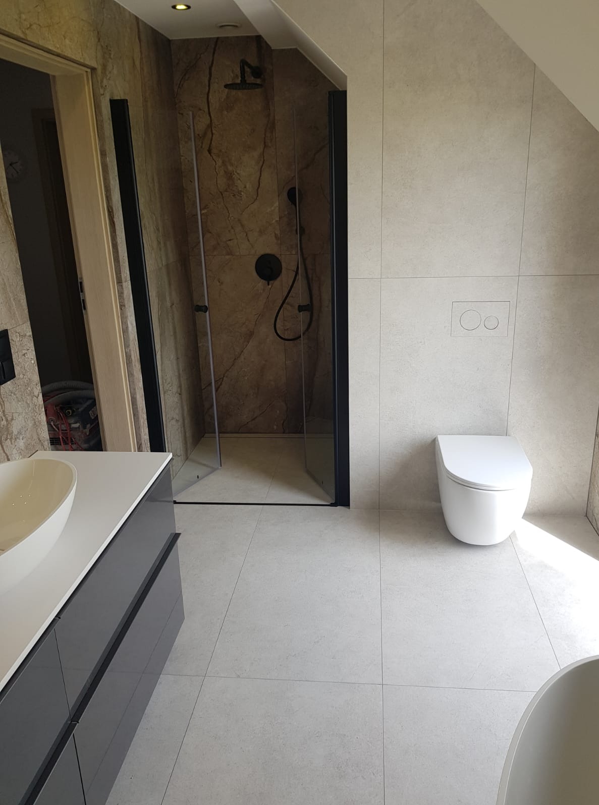Nowoczesna łazienka po remoncie: kabina prysznicowa typu walk-in z czarnymi profilami, podwieszana toaleta i umywalka nablatowa na szafce w odcieniach szarości.