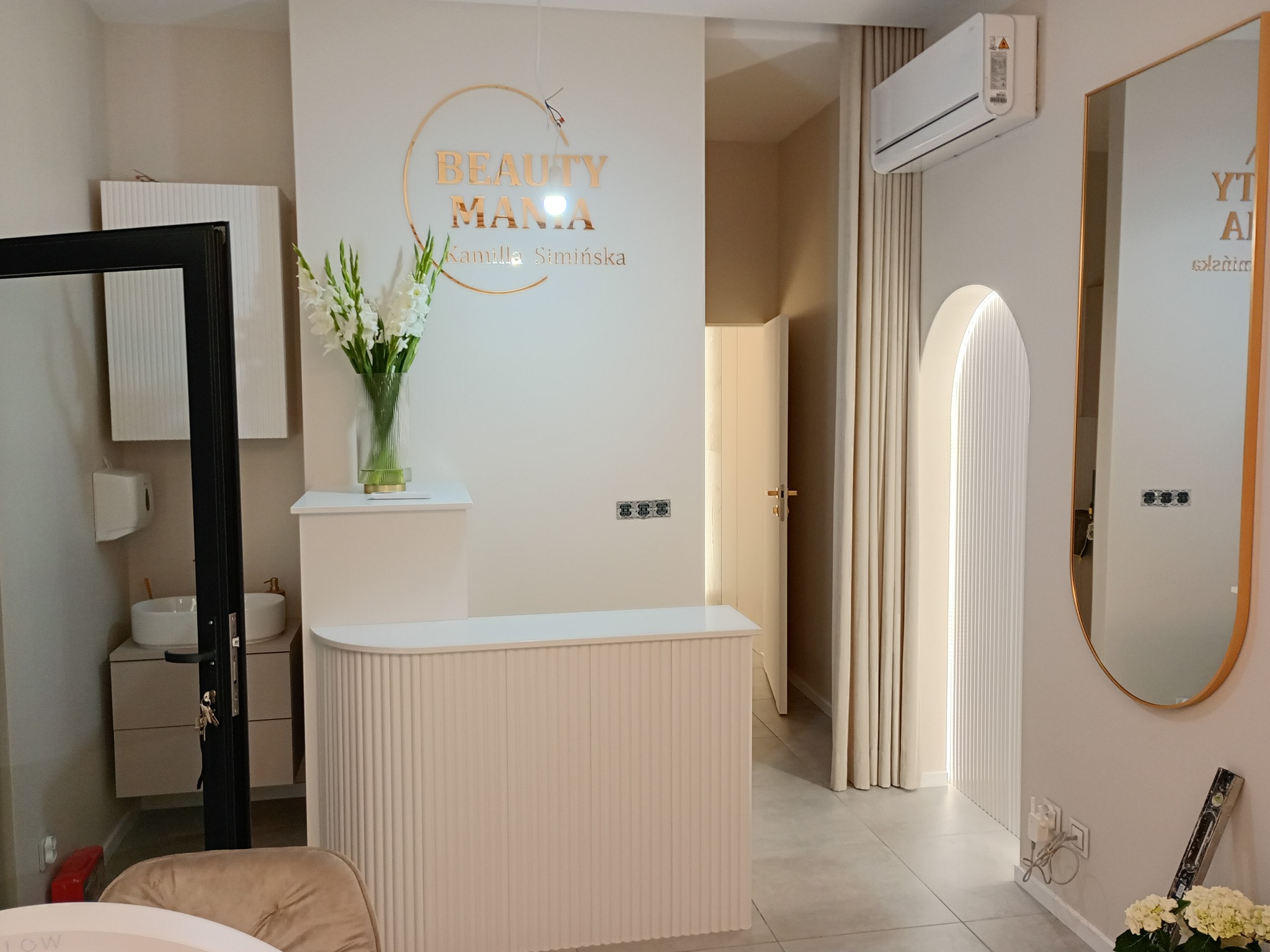 Jasne wnętrze salonu kosmetycznego z recepcją, kwiatami i złotym logo 'Beauty Mania'. Minimalistyczny design z elementami dekoracyjnymi.