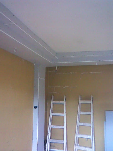 Montaż sufitu podwieszanego z płyt karton gips na metalowym stelażu, widoczne dwie drewniane drabiny opierające się o ścianę w pomarańczowym odcieniu.