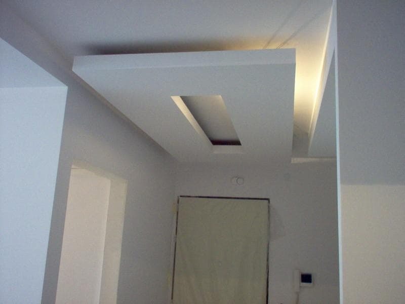 Nowoczesny sufit podwieszany z prostokątną wnęką oświetleniową, białe ściany w tle, widoczne drzwi zabezpieczone folią malarską.