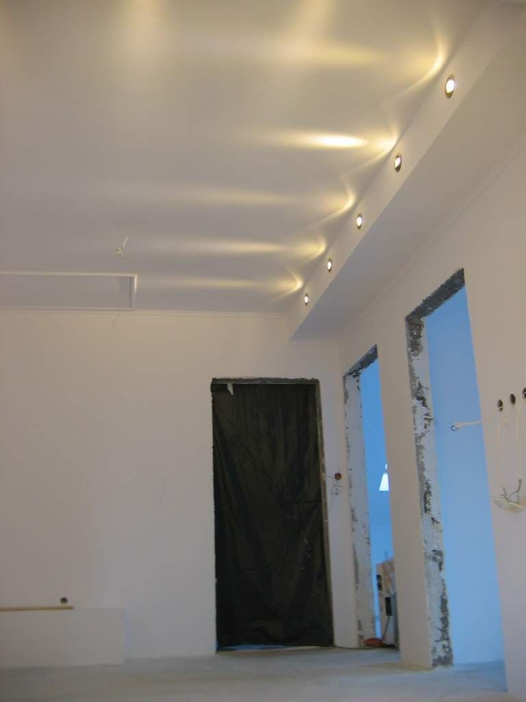 Białe wnętrze w trakcie prac wykończeniowych, widoczne przygotowane otwory drzwiowe, sufit z zamontowanym oświetleniem punktowym i fragment ściany z instalacją elektryczną.