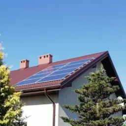 Panele słoneczne na brązowym dachu domu jednorodzinnego z kominami, widoczne z poziomu gruntu, otoczone zielenią drzew iglastych na tle błękitnego nieba.