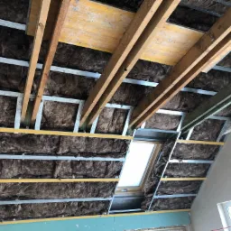 Poddasze w trakcie ocieplania wełną mineralną, widoczne metalowe profile, okno dachowe i fragment płyty gipsowo-kartonowej na ścianie kolankowej.