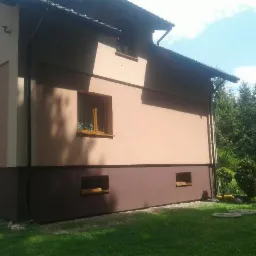 Dwukolorowa elewacja domu jednorodzinnego z drewnianą stolarką okienną, widok z zewnątrz na trawnik i drzewa w tle.