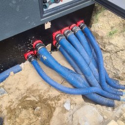 VK ELEKTRO SPÓŁKA Z OGRANICZONĄ ODPOWIEDZIALNOŚCIĄ - Instalacja kabli w karbowanych rurach z czerwonymi złączkami, wychodzących ze skrzynki elektrycznej. Widoczne narzędzia i ziemia. Ujęcie z dołu.