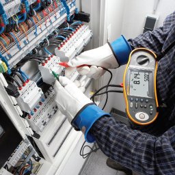 VK ELEKTRO SPÓŁKA Z OGRANICZONĄ ODPOWIEDZIALNOŚCIĄ - Elektryk w rękawicach ochronnych mierzy parametry w rozdzielnicy elektrycznej za pomocą multimetru. Widoczne przewody i zabezpieczenia.