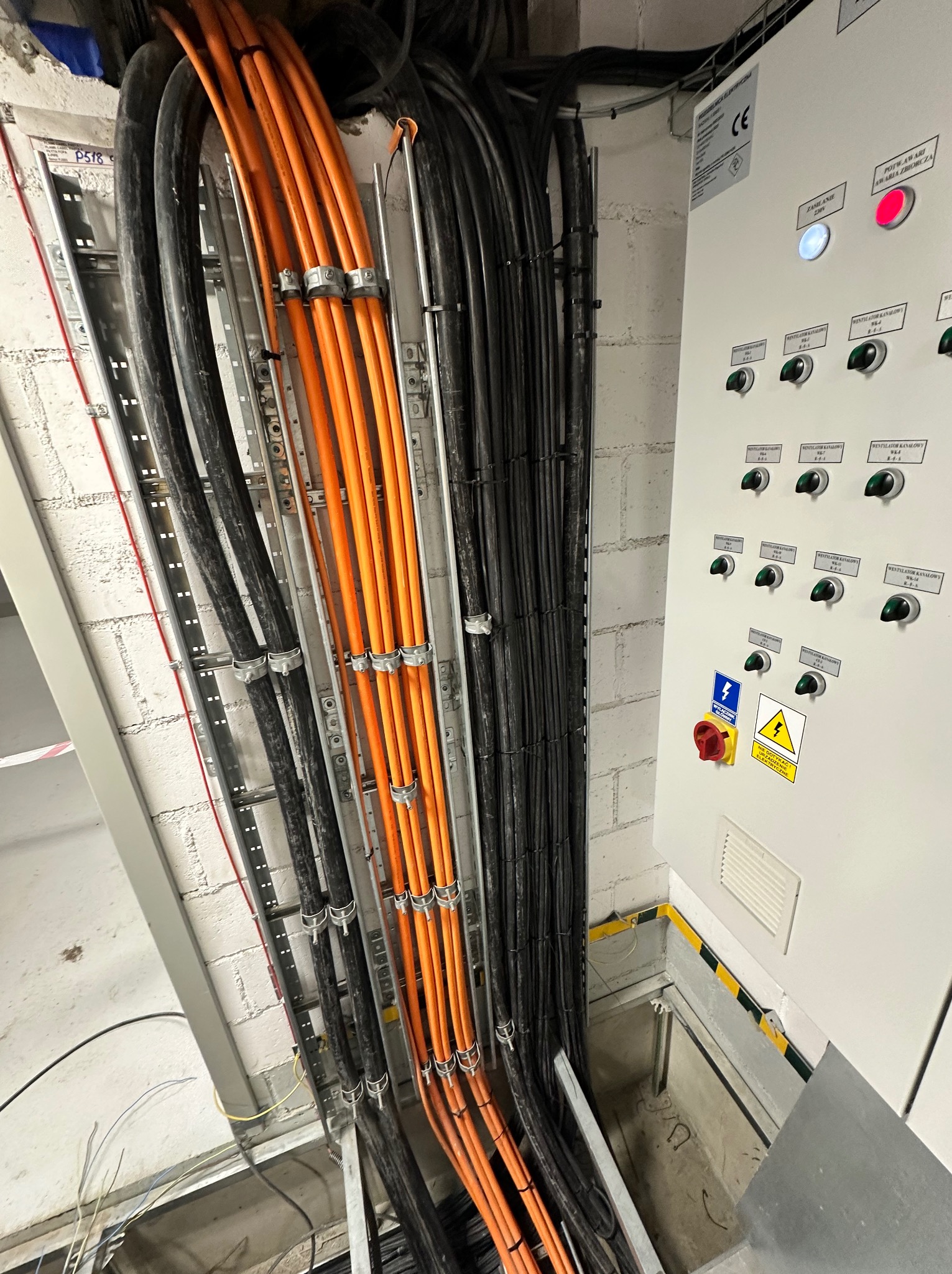 Kable elektryczne w korytach, ułożone równolegle, prowadzone wzdłuż ściany z cegły i szafy sterowniczej z przełącznikami i lampkami kontrolnymi.