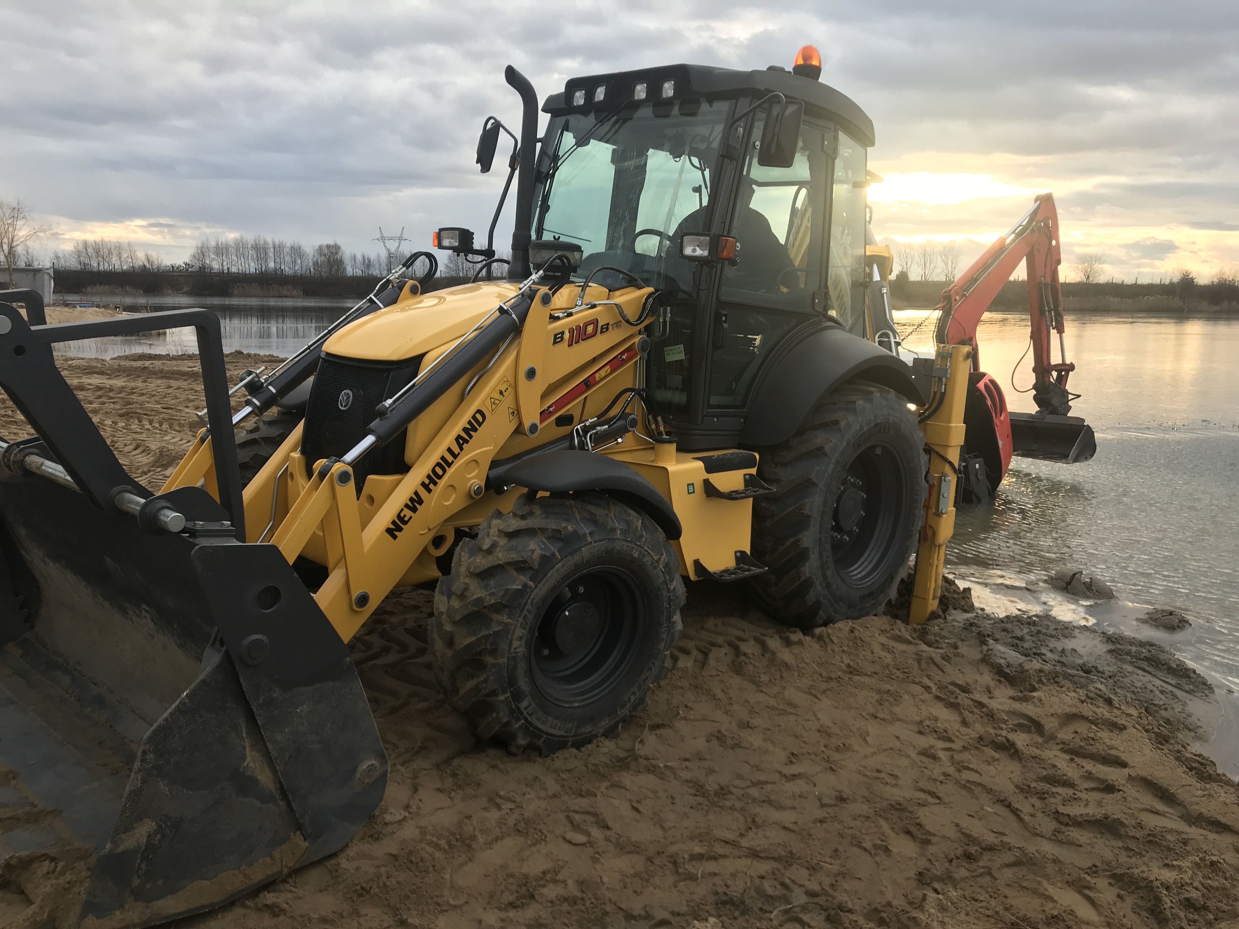 Żółta koparko-ładowarka New Holland B110B TC stoi na piaszczystym brzegu, częściowo zanurzona w wodzie, z widocznym zbiornikiem wodnym i zachodzącym słońcem w tle.