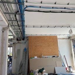ELCOM - INSTALACJE ELEKTRYCZNE - Wnętrze w trakcie remontu z widoczną instalacją elektryczną: kable w korytach i mocowane do sufitu, płyty gipsowo-kartonowe oparte o ścianę, okno przesuwne i pracownik w tle.