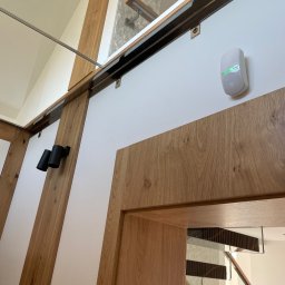 ELCOM - INSTALACJE ELEKTRYCZNE - Czujnik alarmowy z zieloną diodą zamontowany na białej ścianie w nowoczesnym wnętrzu z elementami drewnianymi i szklanymi.