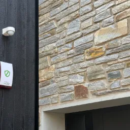 Zewnętrzny czujnik ruchu i sygnalizator alarmowy firmy Loxone zamontowane na elewacji z opalanego drewna i kamienia.