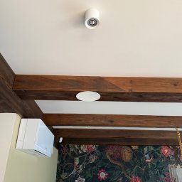 ELCOM - INSTALACJE ELEKTRYCZNE - Wnętrze z drewnianymi belkami sufitowymi, klimatyzatorem ściennym i dwoma okrągłymi, białymi czujnikami na suficie. Na ścianie wzorzysta tapeta.