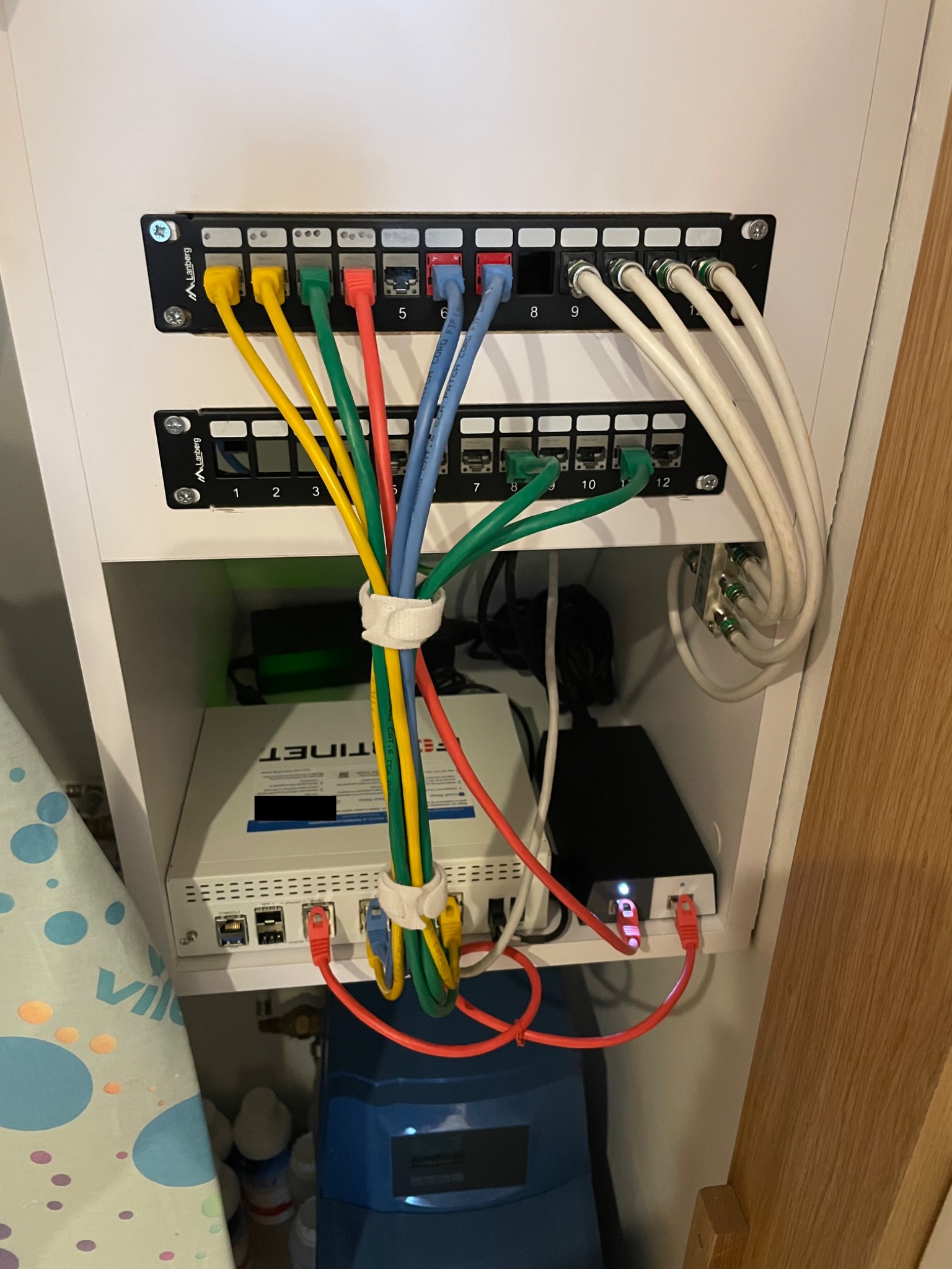 Panel krosowniczy Lanberg z podłączonymi kablami Ethernet w kolorach żółtym, zielonym, czerwonym, niebieskim i białym, zamontowany w białej szafce, poniżej router TINET i czarny zasilacz.