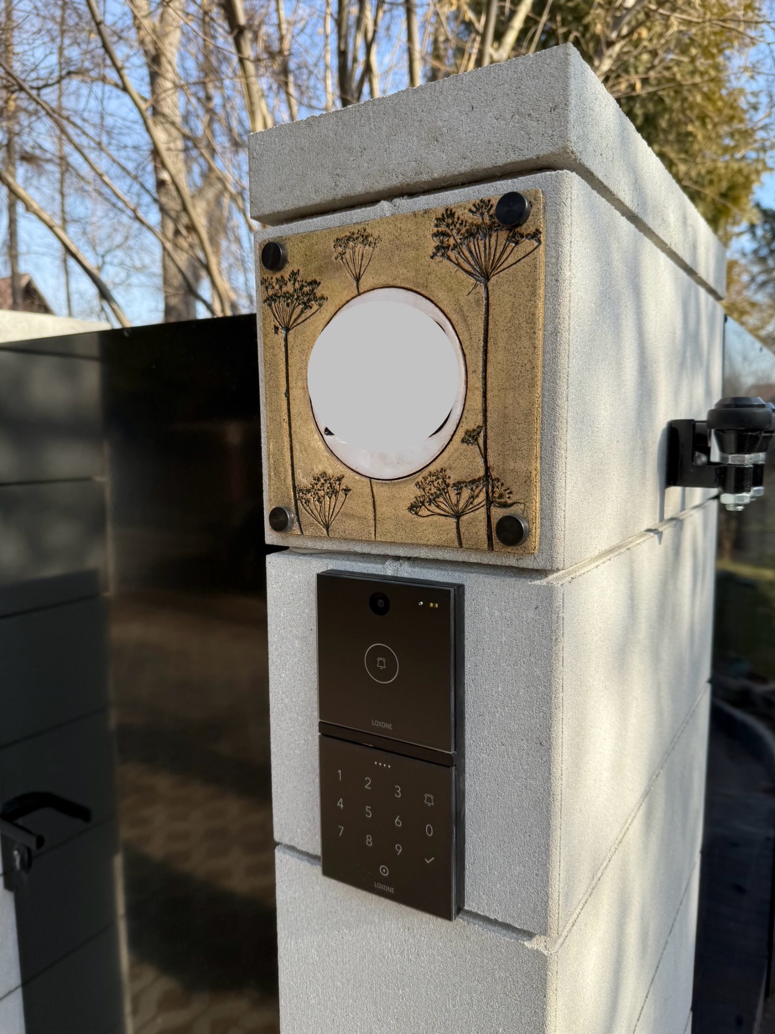 Brama wjazdowa z wideodomofonem Loxone i ozdobnym panelem z motywem roślinnym, zamontowane na betonowym słupku.