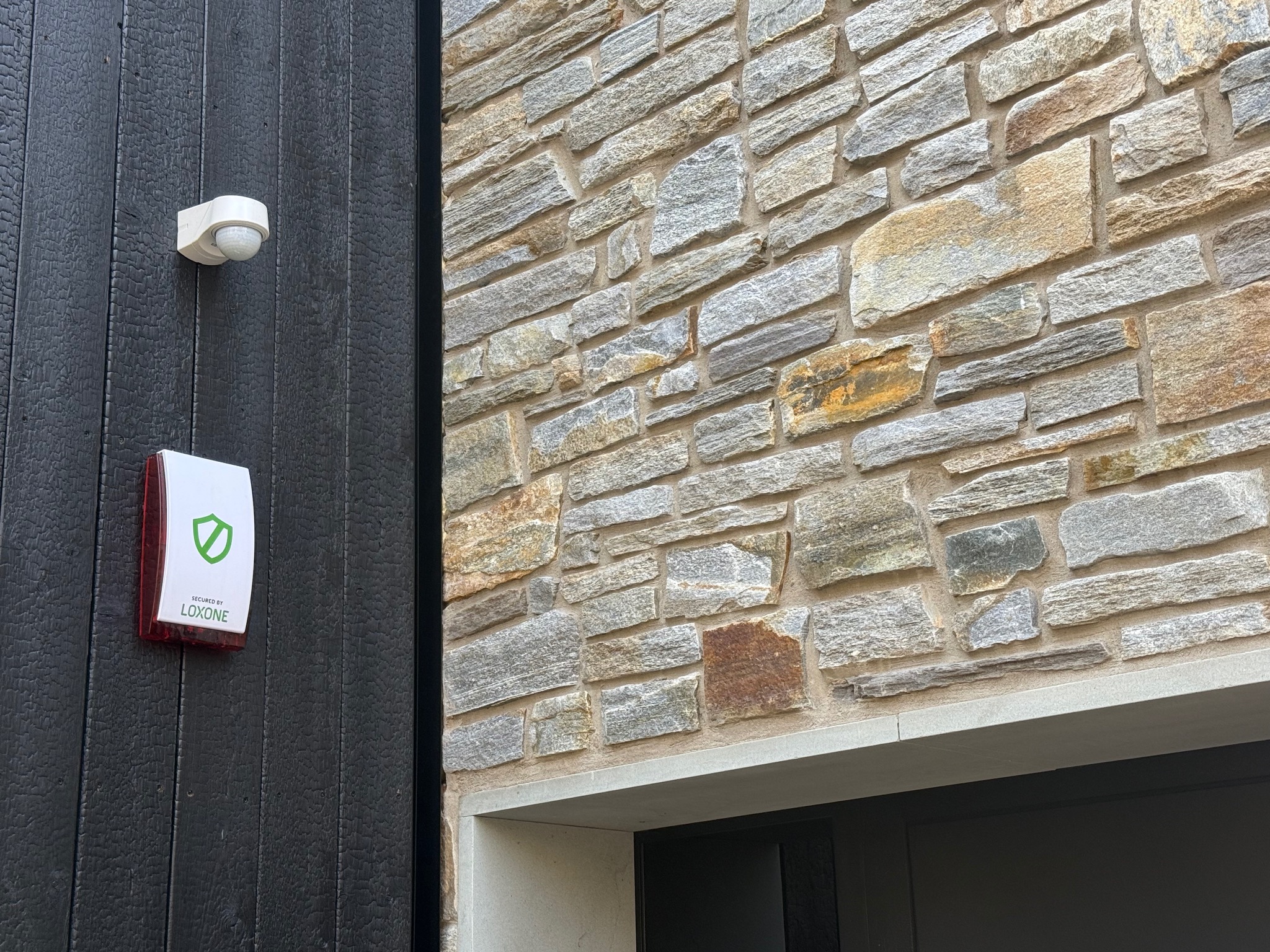 Zewnętrzny czujnik ruchu i sygnalizator alarmowy firmy Loxone zamontowane na elewacji z opalanego drewna i kamienia.