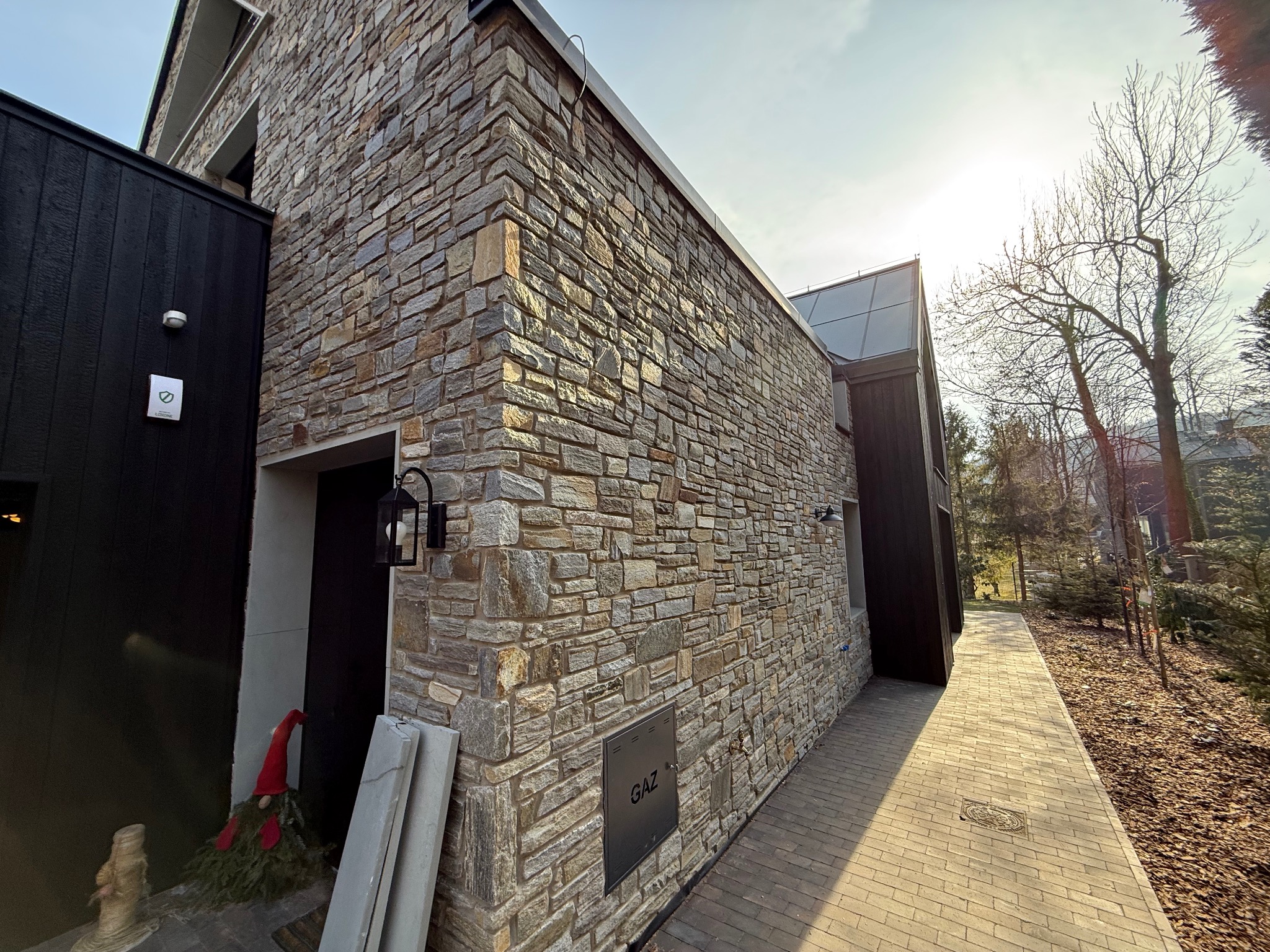 Fragment elewacji budynku wykończonej kamieniem naturalnym, z widoczną czarną skrzynką gazową i oświetleniem zewnętrznym, oraz ścieżką z kostki brukowej. Obok wejścia stoi dekoracja świąteczna.