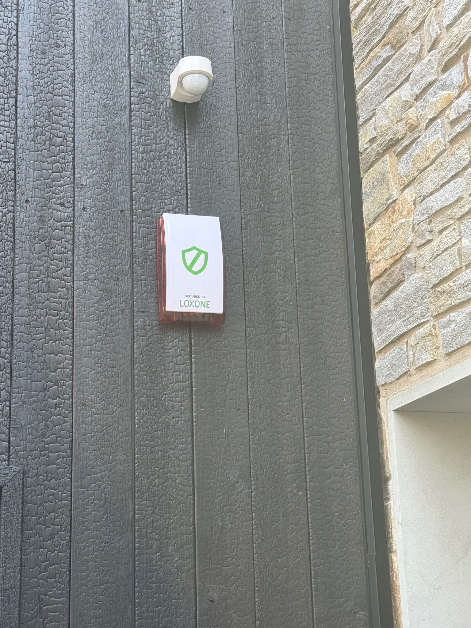 Zewnętrzny czujnik ruchu i sygnalizator alarmowy firmy Loxone zamontowany na elewacji z ciemnego drewna, obok ściany z kamienia.