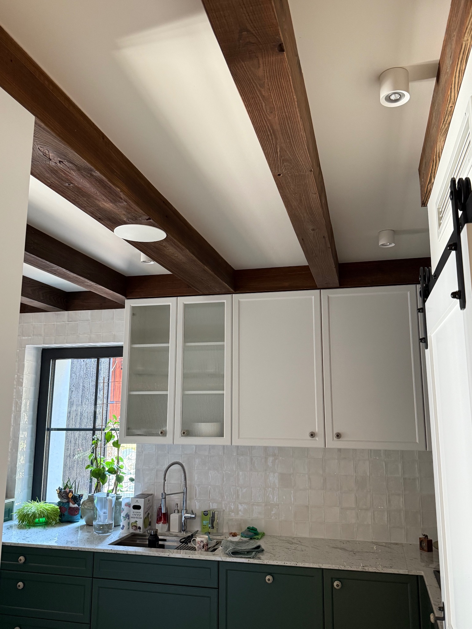 Kuchnia z ciemnozielonymi szafkami dolnymi, białymi szafkami wiszącymi z przeszklonymi frontami, białymi płytkami na ścianie i drewnianymi belkami stropowymi