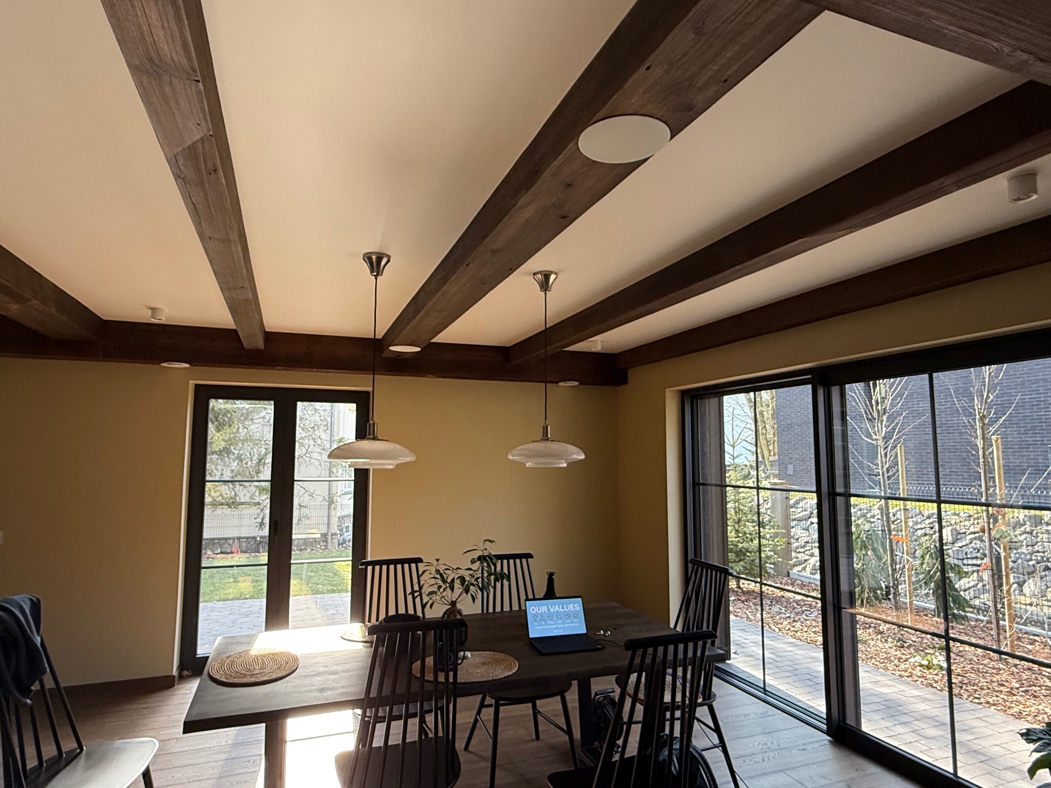 Jasne wnętrze jadalni z drewnianymi belkami stropowymi i dużymi oknami przesuwnymi, widok na ogród, laptop na stole.