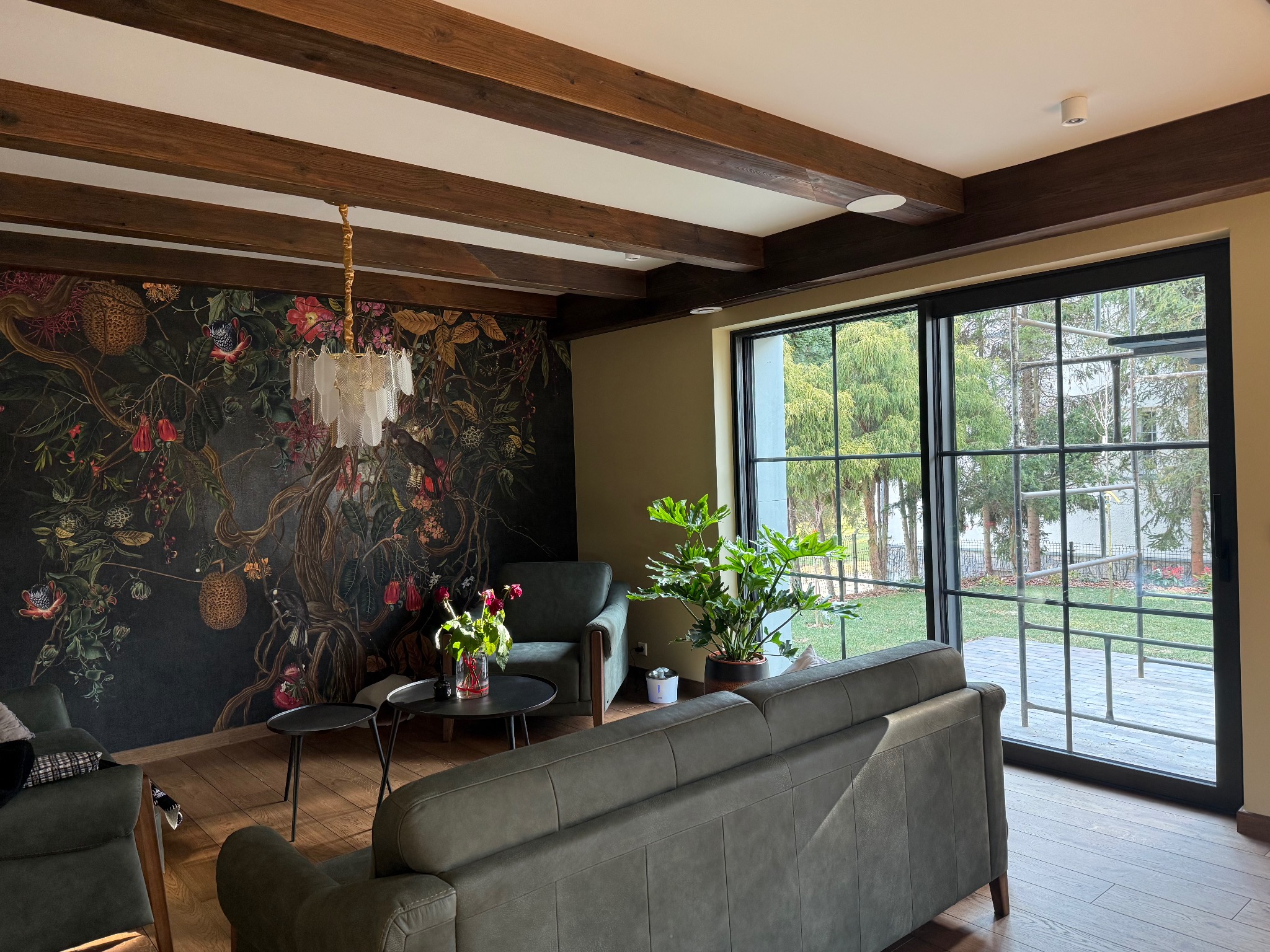 Jasny salon z drewnianymi belkami stropowymi, ozdobną tapetą z motywami roślinnymi, nowoczesnym oświetleniem i dużym oknem z widokiem na ogród.