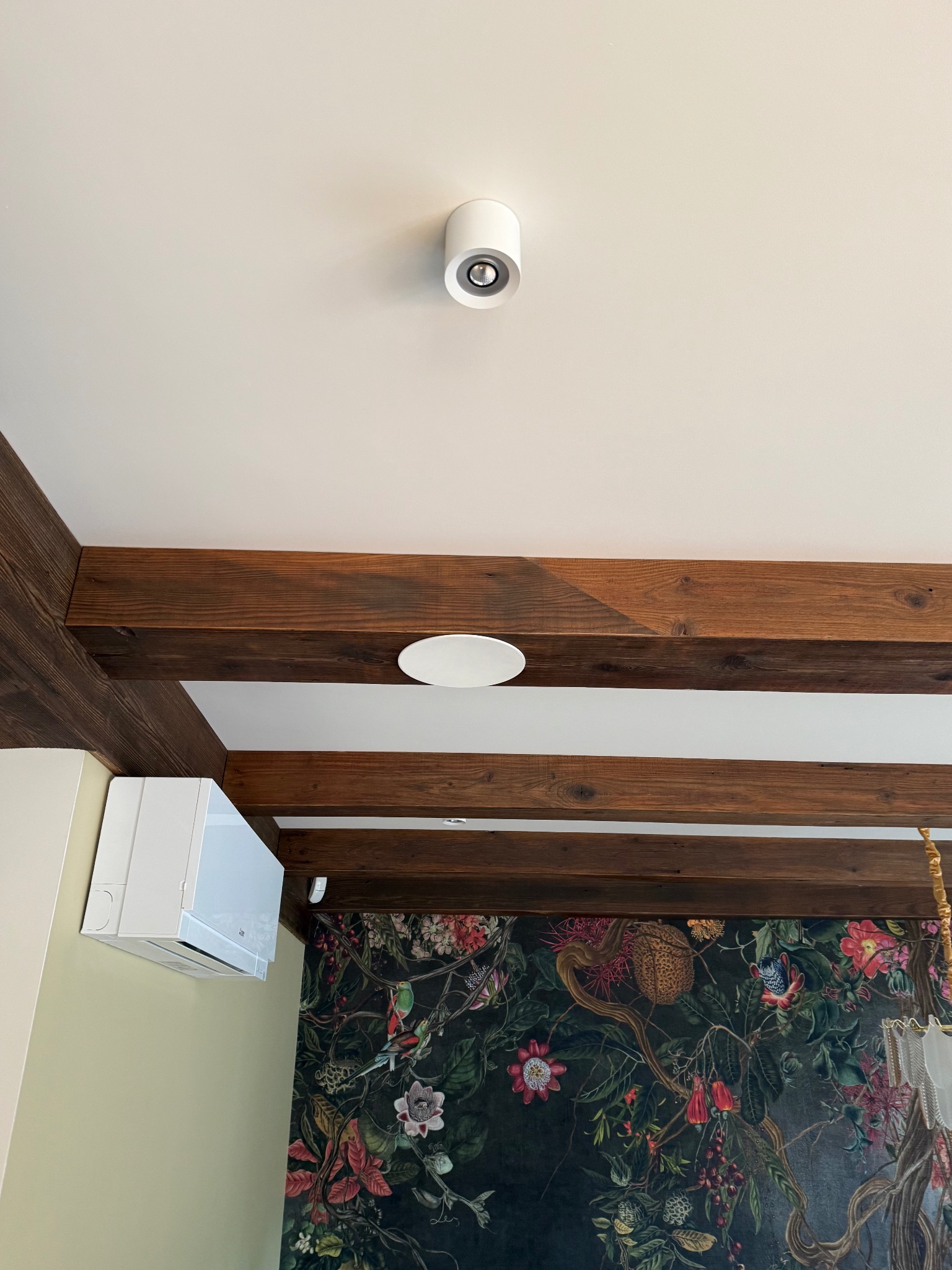 Wnętrze z drewnianymi belkami sufitowymi, klimatyzatorem ściennym i dwoma okrągłymi, białymi czujnikami na suficie. Na ścianie wzorzysta tapeta.