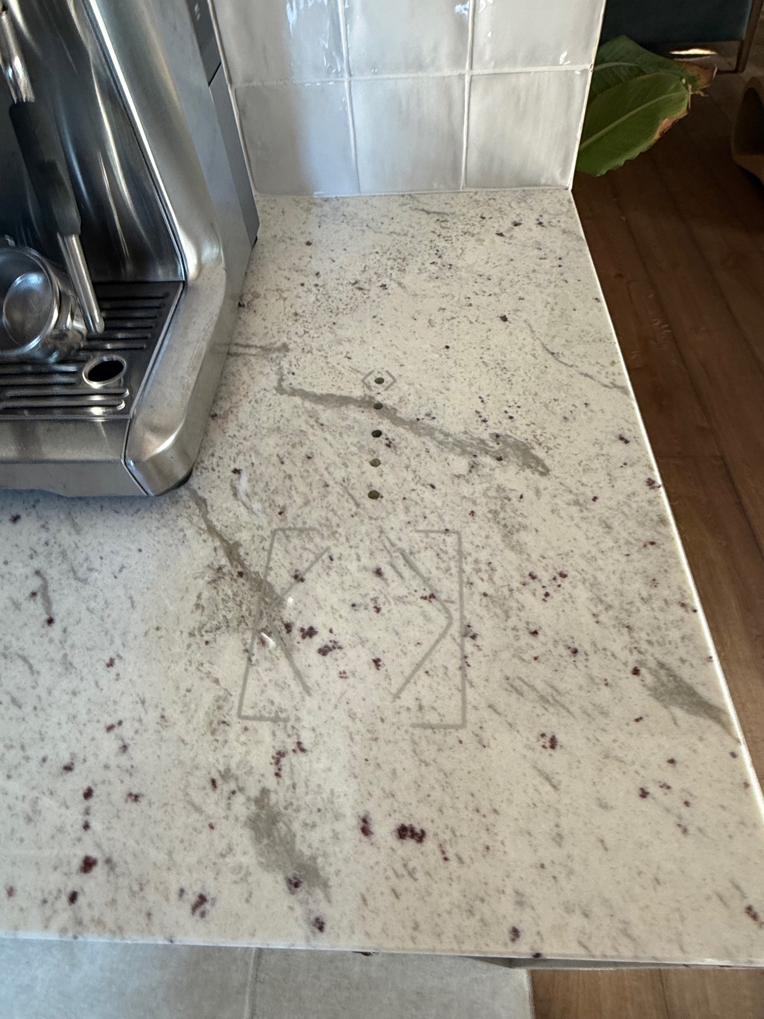 Fragment blatu kuchennego z jasnego, nakrapianego granitu z wygrawerowanym symbolem strzałek w dół i na boki, obok ekspresu do kawy i płytek ściennych.