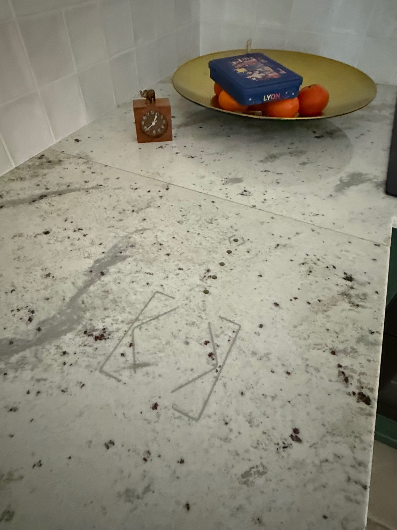 Fragment blatu kuchennego z elementami dekoracyjnymi: drewniany zegar z figurką słonia, złota misa z owocami i pudełkiem, oraz zarys prostokątnej obudowy po urządzeniu.