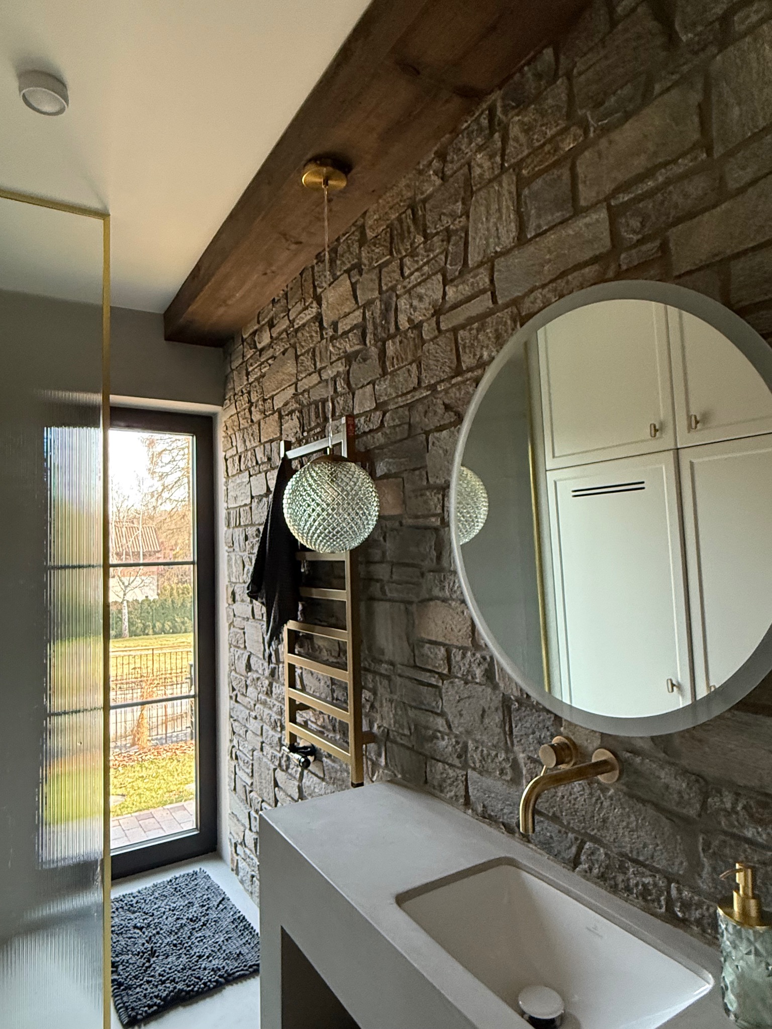 Nowoczesna łazienka w Nowym Wiśniczu z kamienną ścianą, okrągłym lustrem, szarą umywalką i złotymi elementami wykończenia, w tym grzejnik drabinkowy i bateria umywalkowa.