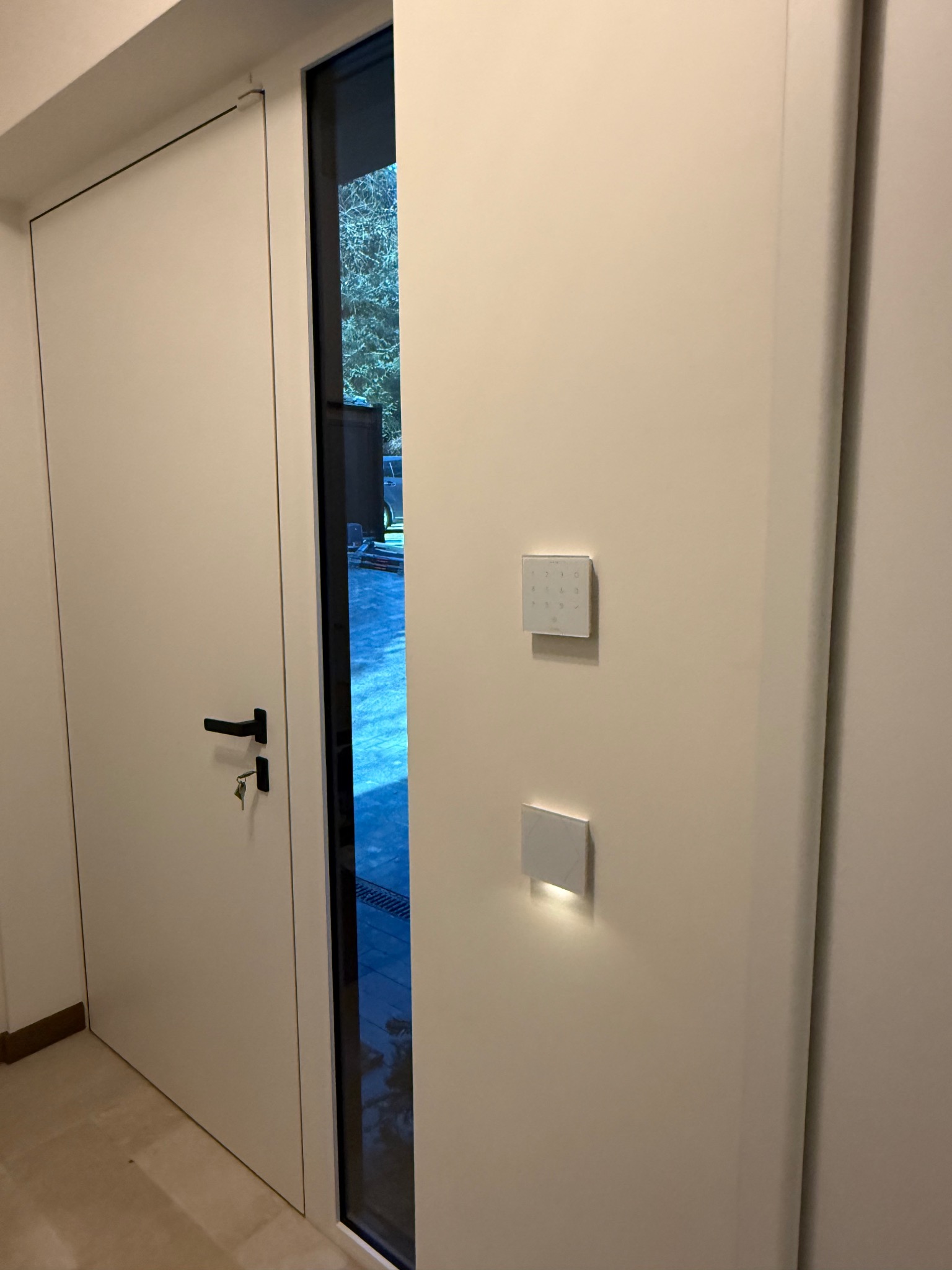 Białe panele sterowania systemem inteligentnego domu zamontowane na ścianie obok drzwi wejściowych z czarną klamką i kluczem w zamku.