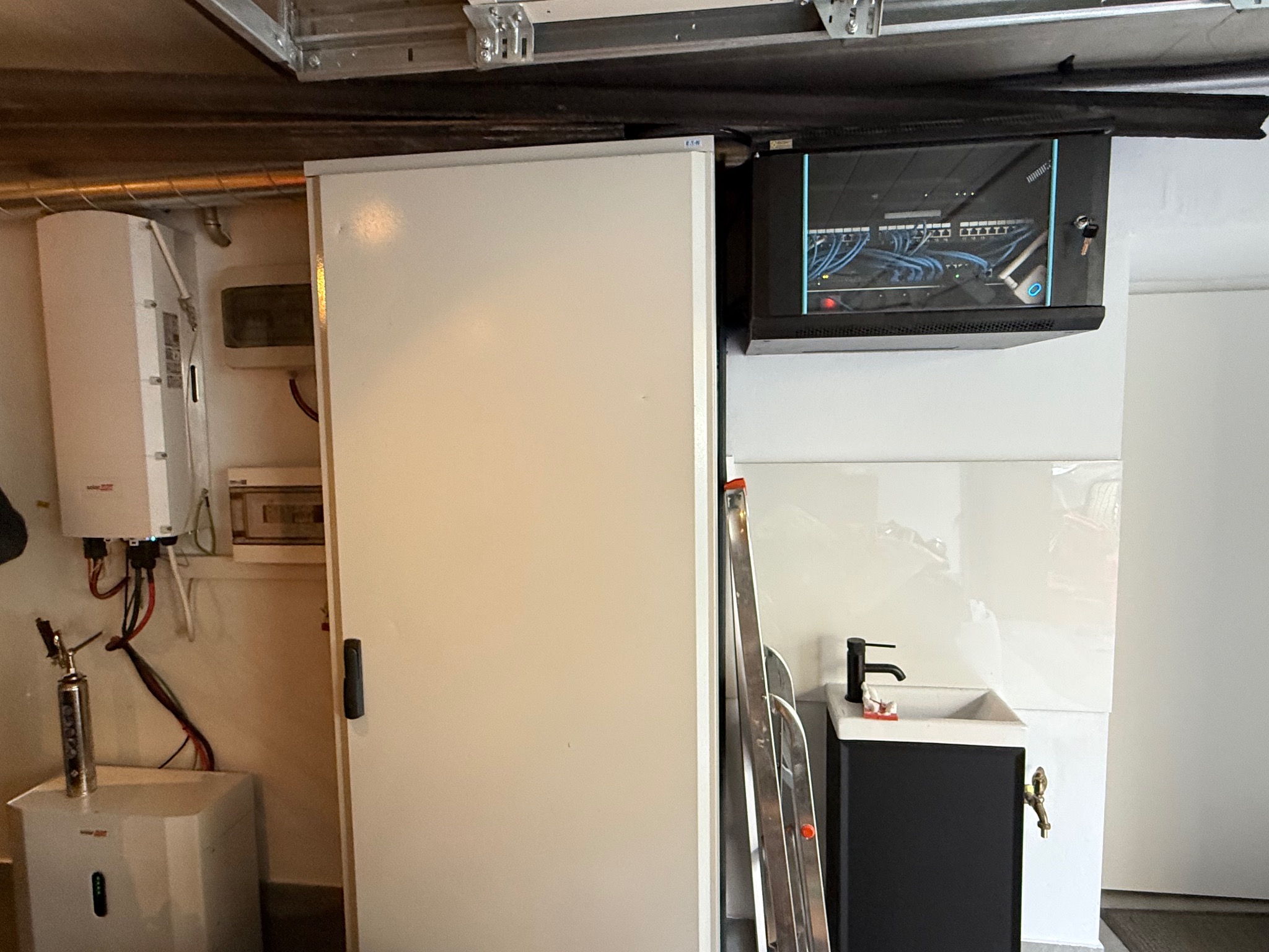 Szafa rack z okablowaniem, panel sterowania, biała szafka z drzwiami, drabina i umywalka w pomieszczeniu technicznym.