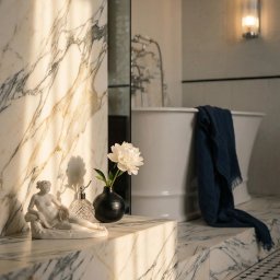 SUN HOUSE DESIGN SPÓŁKA Z OGRANICZONĄ ODPOWIEDZIALNOŚCIĄ - Elegancka łazienka z marmurowymi elementami: wanna wolnostojąca, mozaika na podłodze, rzeźba i kwiat w wazonie w promieniach słońca. Lampa ścienna w tle.