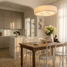 SUN HOUSE DESIGN SPÓŁKA Z OGRANICZONĄ ODPOWIEDZIALNOŚCIĄ - Jasna kuchnia z wyspą i drewnianym stołem. Na blacie wazon z kwiatami, rzeźba kobiety i filiżanka. Nad stołem okrągła lampa. W tle widoczna marmurowa wyspa.