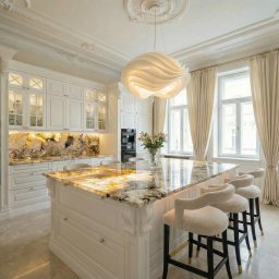 SUN HOUSE DESIGN SPÓŁKA Z OGRANICZONĄ ODPOWIEDZIALNOŚCIĄ - Elegancka, jasna kuchnia z wyspą o marmurowym blacie, ozdobnym oświetleniem i sztukaterią sufitową. Widoczne krzesła barowe i okna z kremowymi zasłonami.