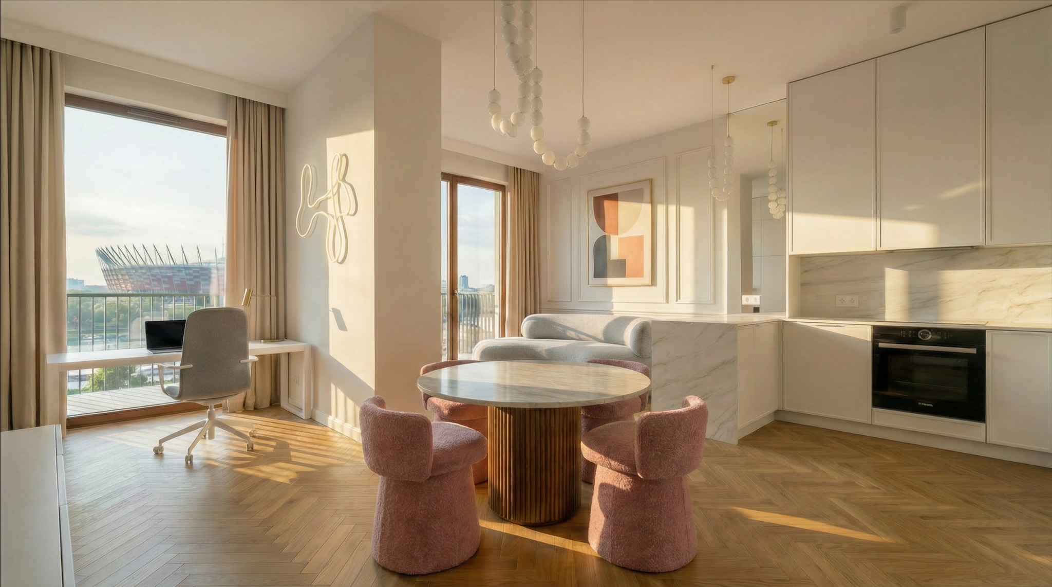Jasne wnętrze mieszkania z widokiem na Stadion Narodowy. Parkiet w jodełkę, okrągły stół z różowymi krzesłami, biała kuchnia i biurko z laptopem przy oknie.