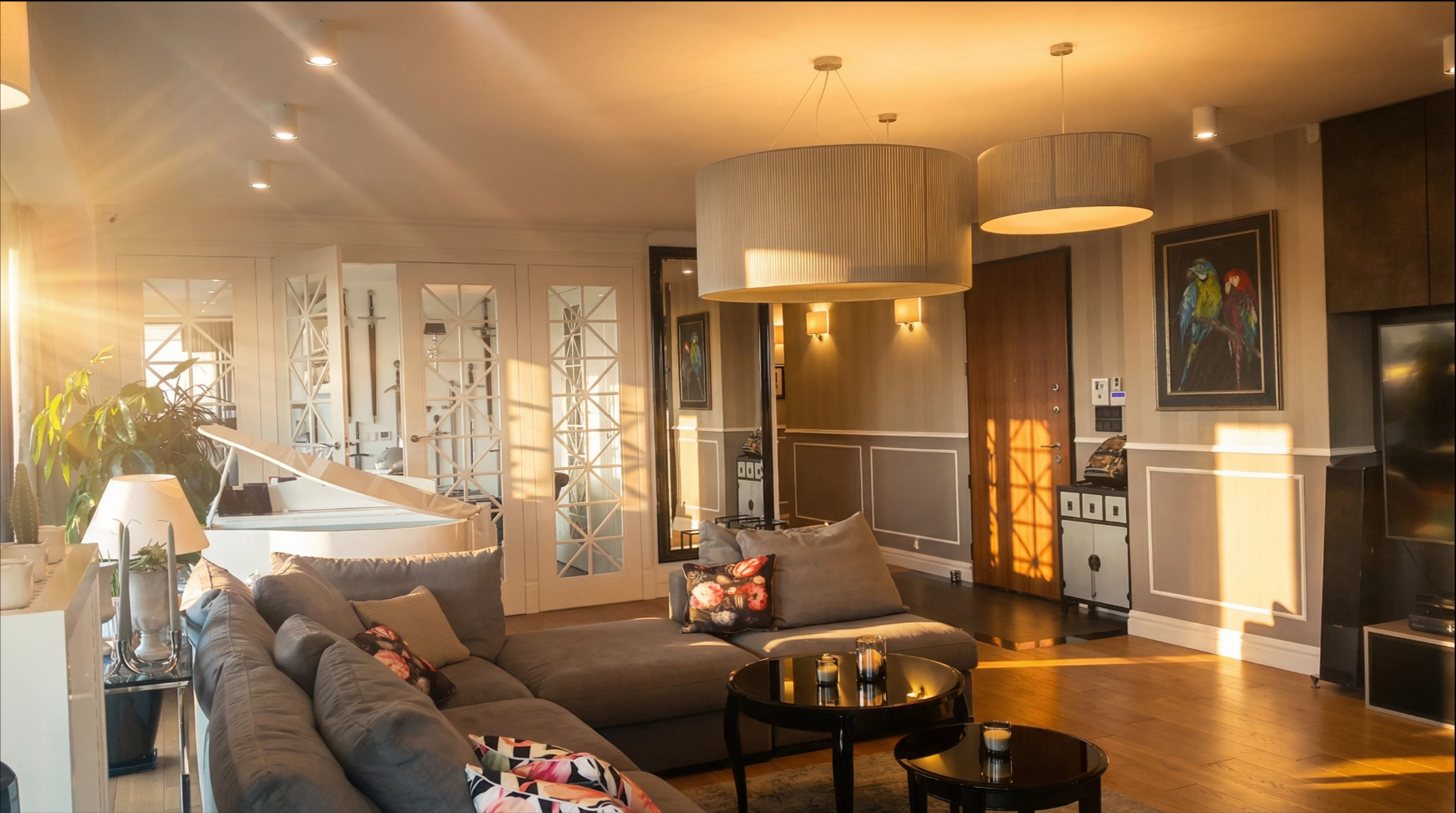 Salon z szarą sofą narożną, białym pianinem i promieniami słońca wpadającymi przez okno. Dwa okrągłe żyrandole nad stolikami kawowymi.