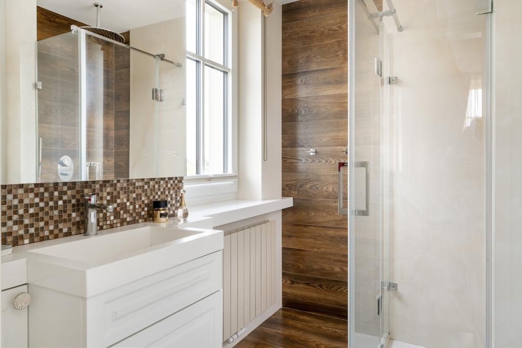 Nowoczesna łazienka z kabiną prysznicową, drewnopodobnymi płytkami na ścianie i podłodze, białą umywalką nablatową z mozaikową ścianą nad nią oraz oknem z białym parapetem.