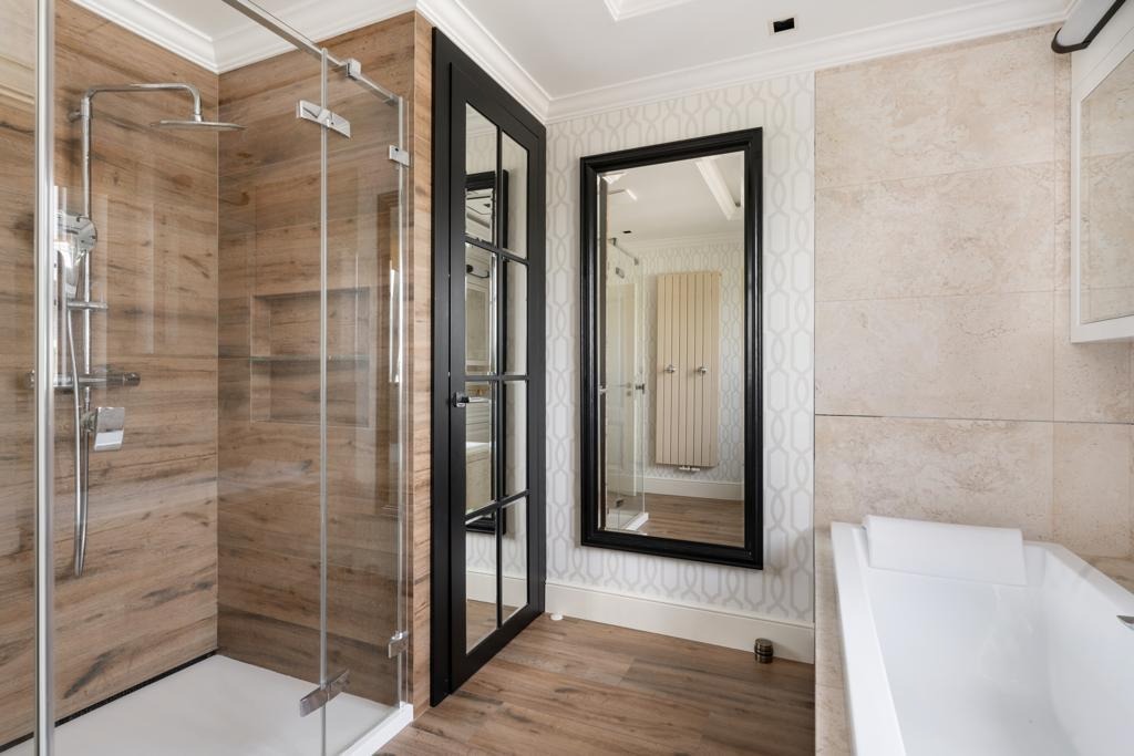 Nowoczesna łazienka w Warszawie z kabiną prysznicową typu walk-in z drewnopodobnymi płytkami, czarną ramą drzwi i lustra, oraz białą wanną.