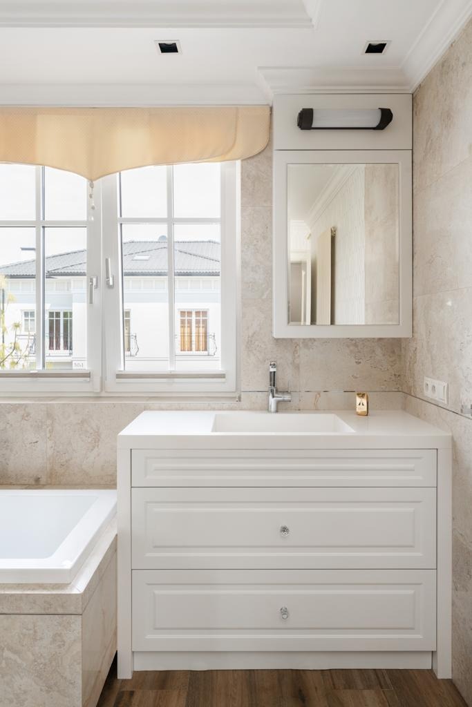 Jasna łazienka z umywalką w zabudowie z szufladami, lustrem w szafce nad umywalką i oknem z widokiem na dom.