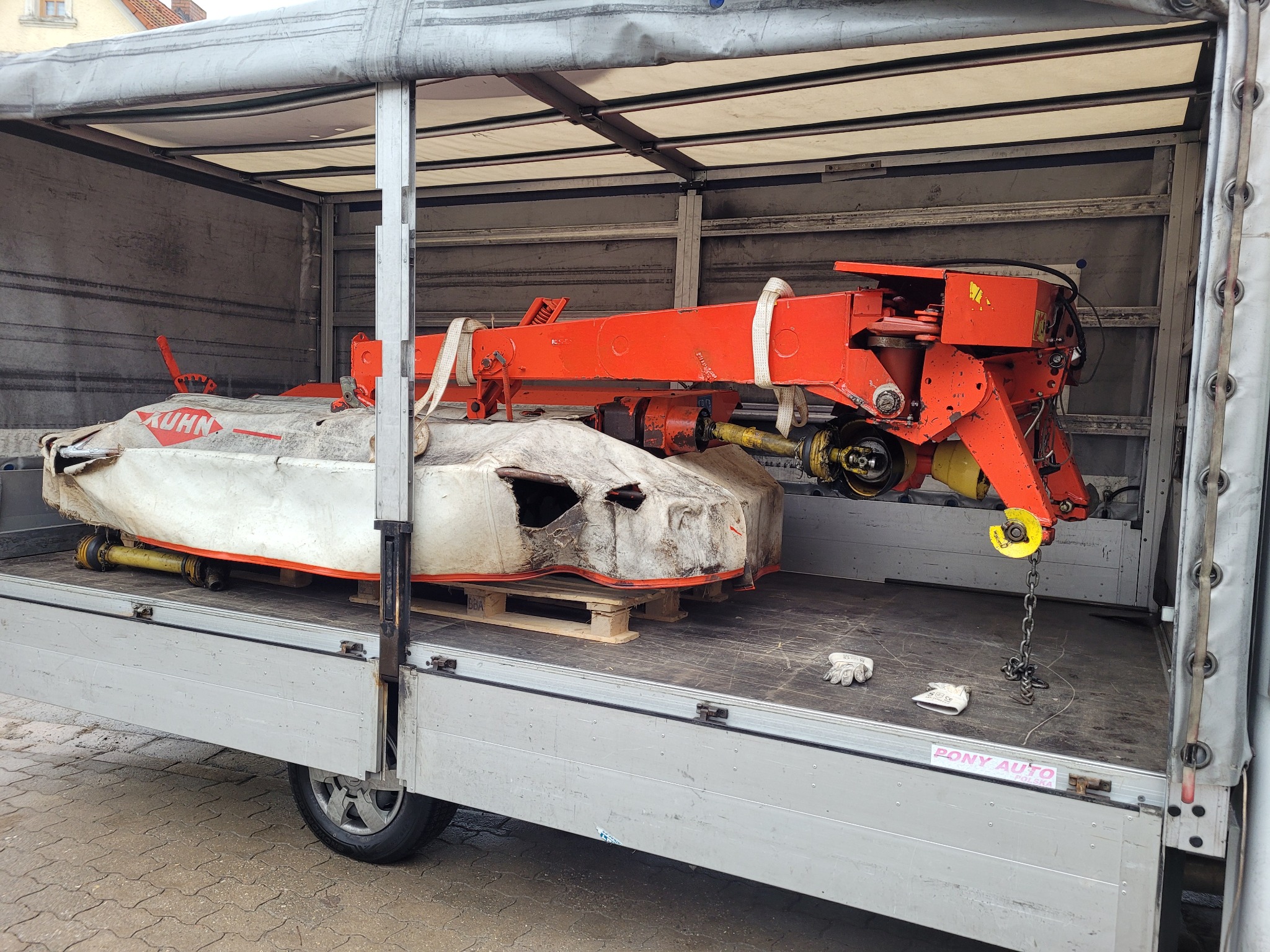 Pomarańczowa kosiarka rolnicza marki Kuhn zabezpieczona pasami w przestrzeni ładunkowej samochodu dostawczego z plandeką, widoczne rękawice robocze i łańcuch.